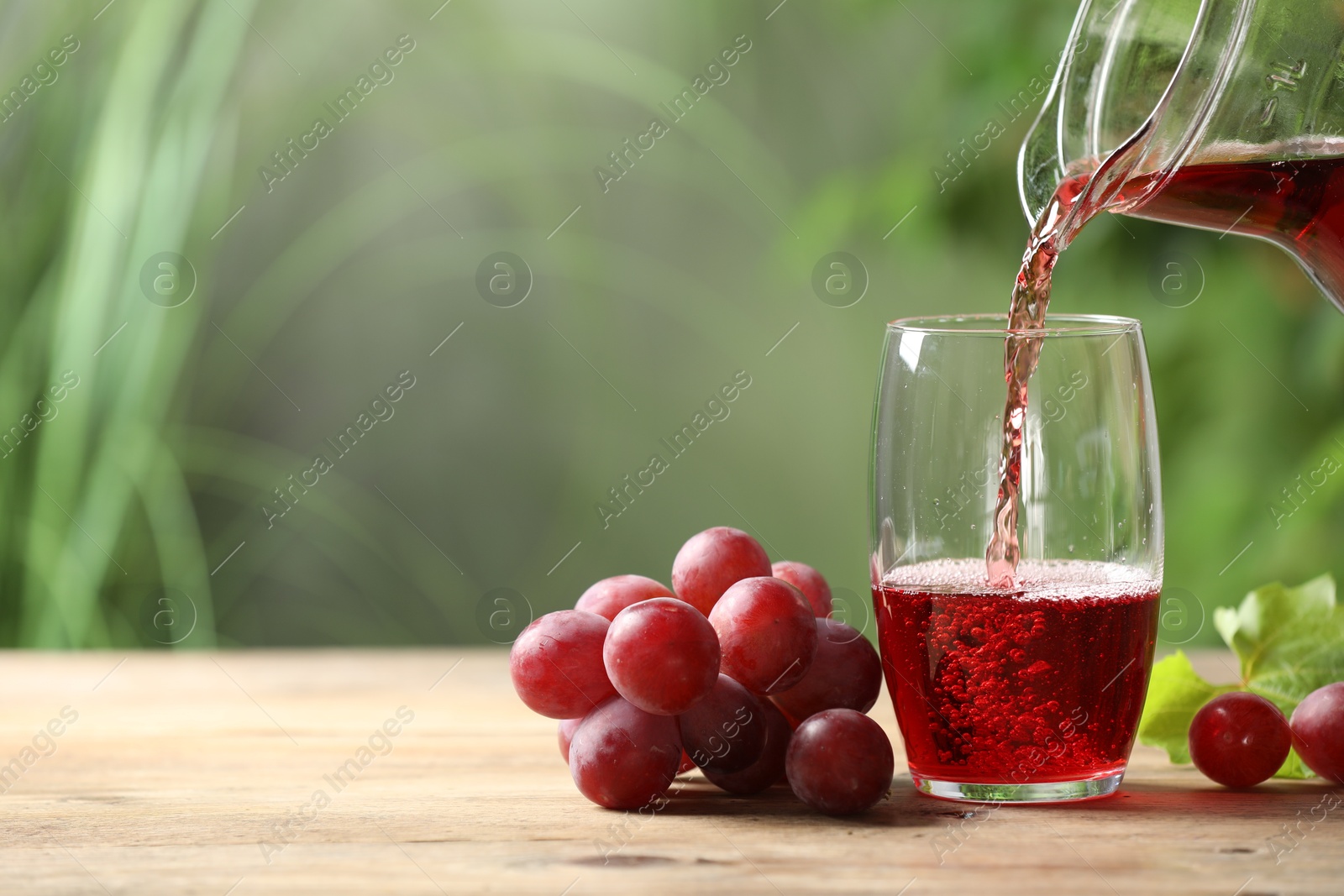 Photo of Pouring tasty grape juice into glass at wooden table outdoors, closeup. Space for text