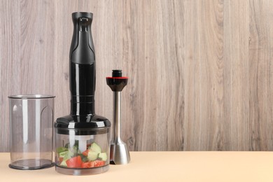 Hand blender kit with fresh vegetables and parsley on beige table against wooden background, space for text