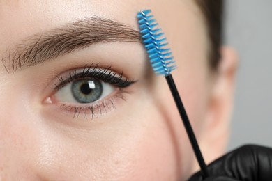 Photo of Beautician brushing young woman's eyebrow on light background, closeup