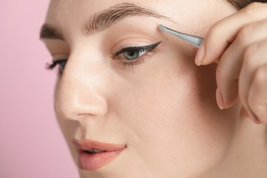 Photo of Young woman plucking eyebrow with tweezers on pink background, closeup