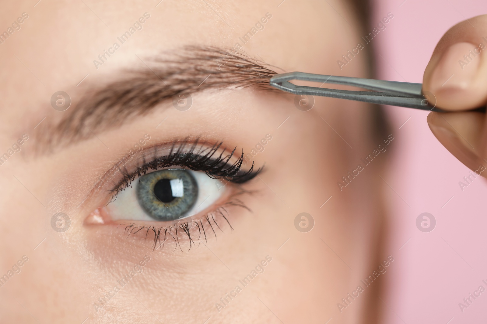 Photo of Young woman plucking eyebrow with tweezers on pink background, closeup