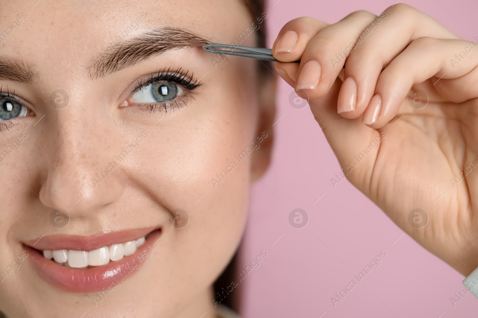 Photo of Young woman plucking eyebrow with tweezers on pink background, closeup