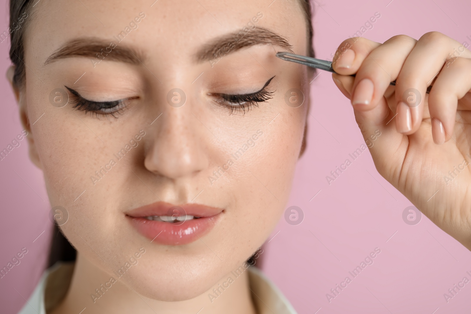 Photo of Young woman plucking eyebrow with tweezers on pink background, closeup