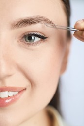Photo of Young woman plucking eyebrow with tweezers on light background, closeup