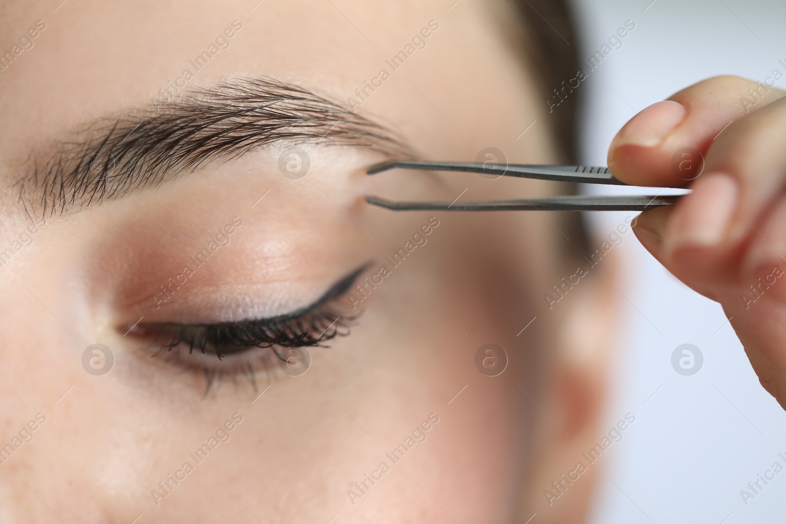 Photo of Young woman plucking eyebrow with tweezers on light background, closeup