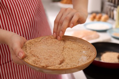 Photo of Woman holding raw schnitzels with breadcrumbs in kitchen, closeup