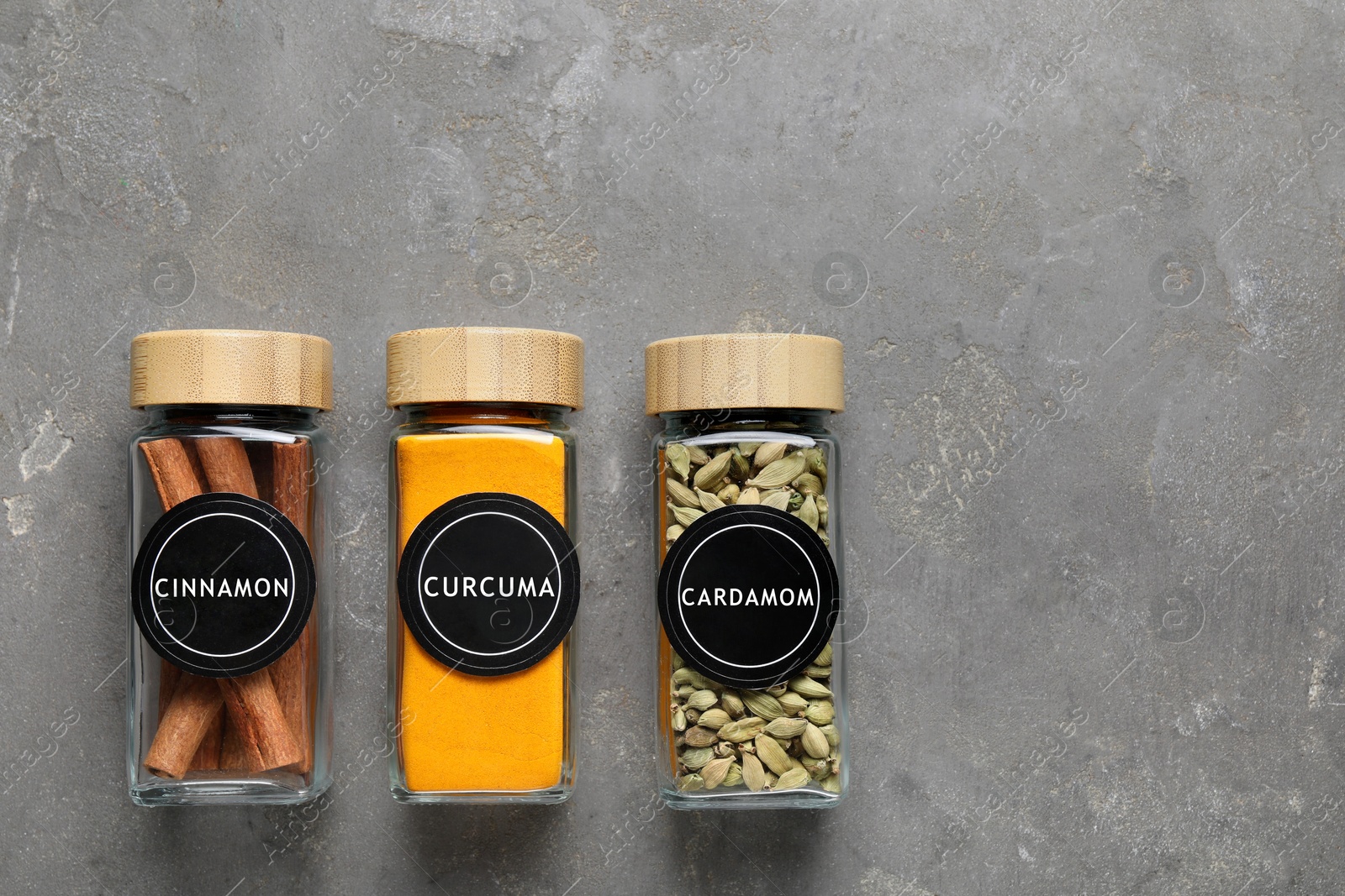 Image of Labelled jars with spices (cinnamon, curcuma and cardamom) on grey table, flat lay. Space for text