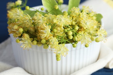 Fresh linden leaves and flowers in bowl on blue wooden table, closeup