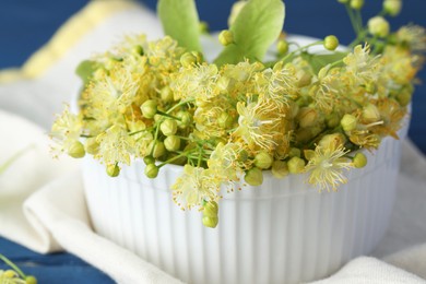 Photo of Fresh linden leaves and flowers in bowl on blue wooden table, closeup