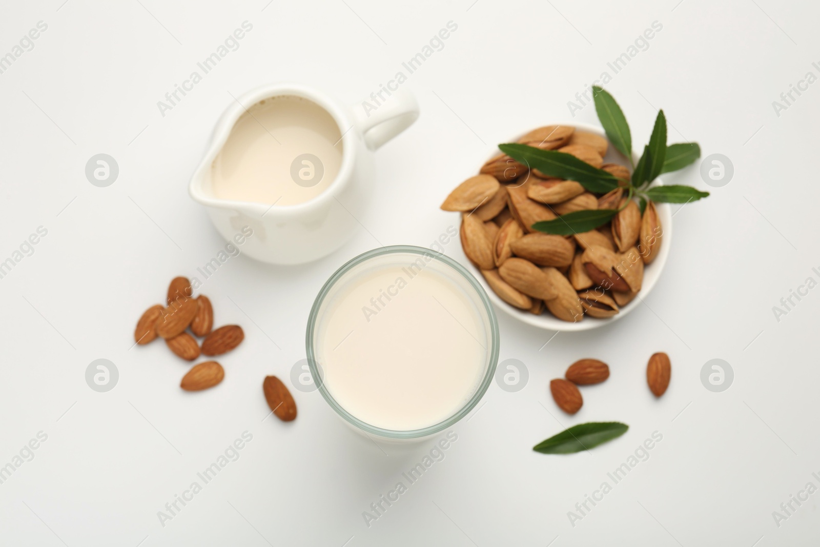 Photo of Fresh almond milk in glass, nuts, green leaves and pitcher on white background, flat lay