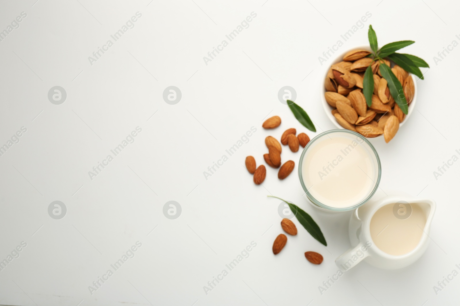 Photo of Fresh almond milk in glass, nuts, green leaves and pitcher on white background, flat lay. Space for text