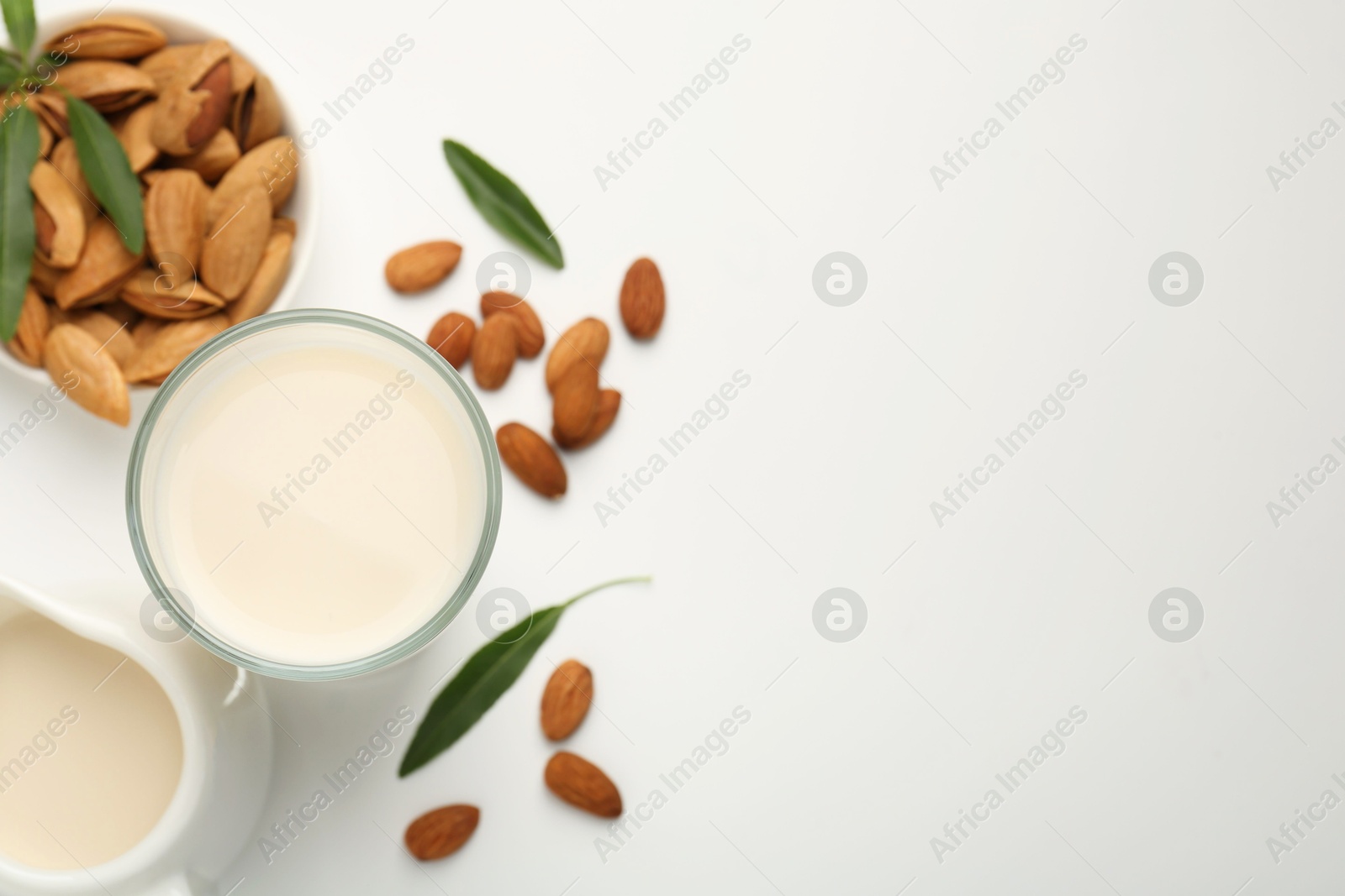 Photo of Fresh almond milk in glass, nuts, green leaves and pitcher on white background, flat lay. Space for text