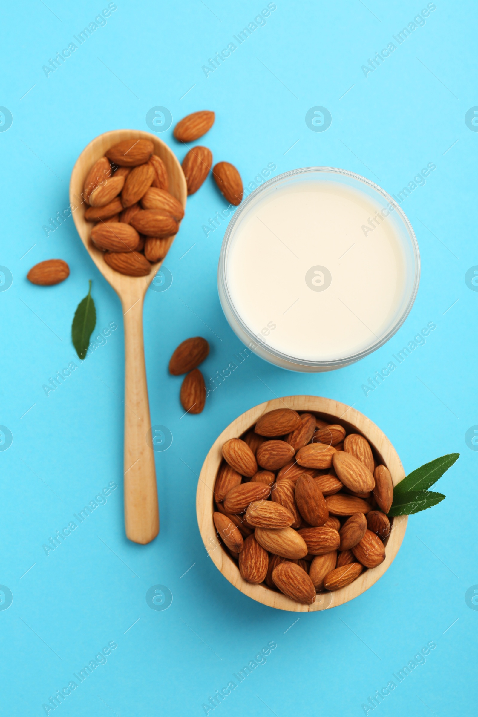 Photo of Fresh almond milk in glass, nuts, spoon and green leaves on light blue background, flat lay