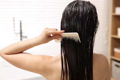 Photo of Woman combing her hair with applied mask in bathroom, back view