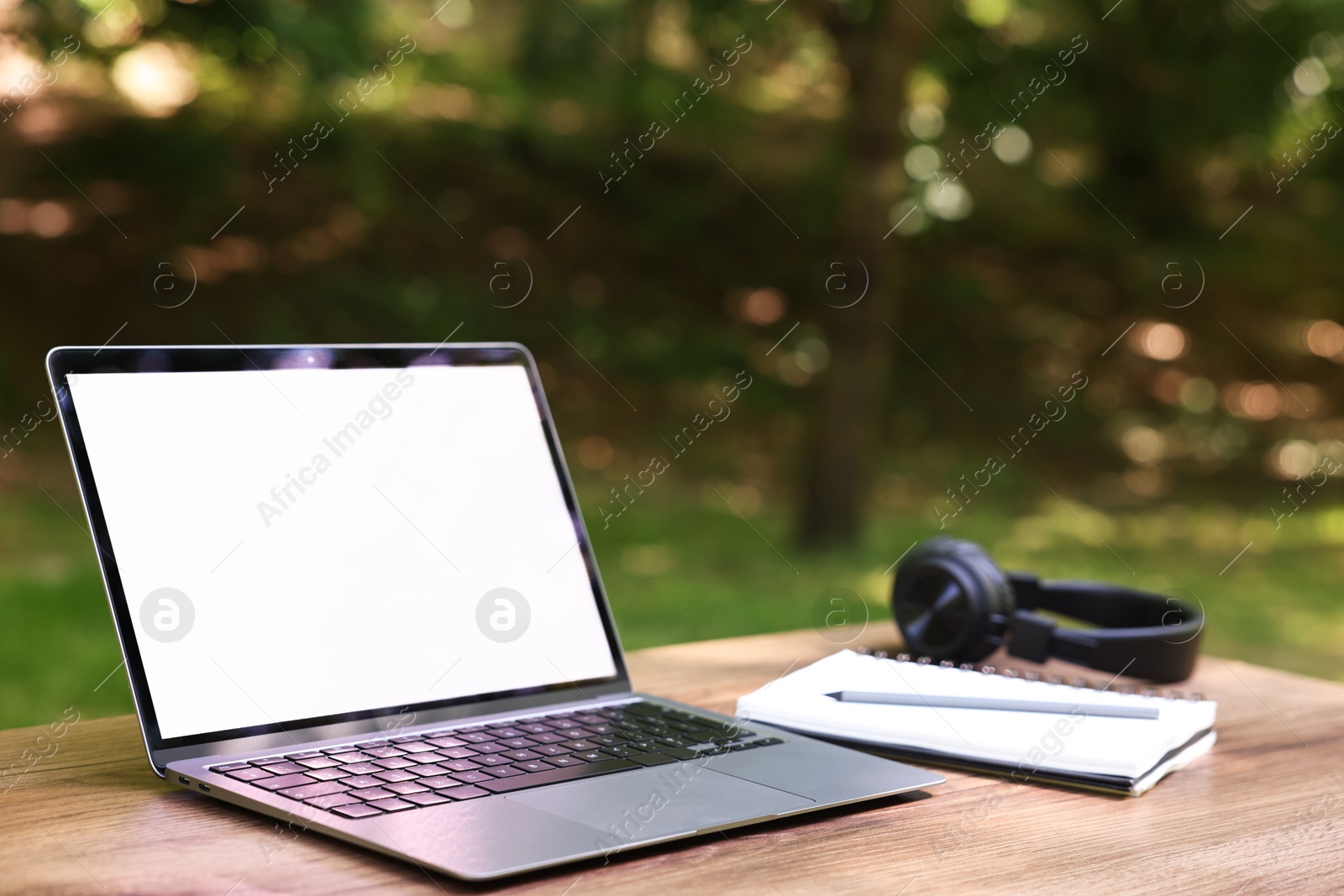 Photo of Laptop, notebook, pen and headphones on wooden table outdoors. Remote work