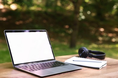 Photo of Laptop, notebook, pen and headphones on wooden table outdoors. Remote work