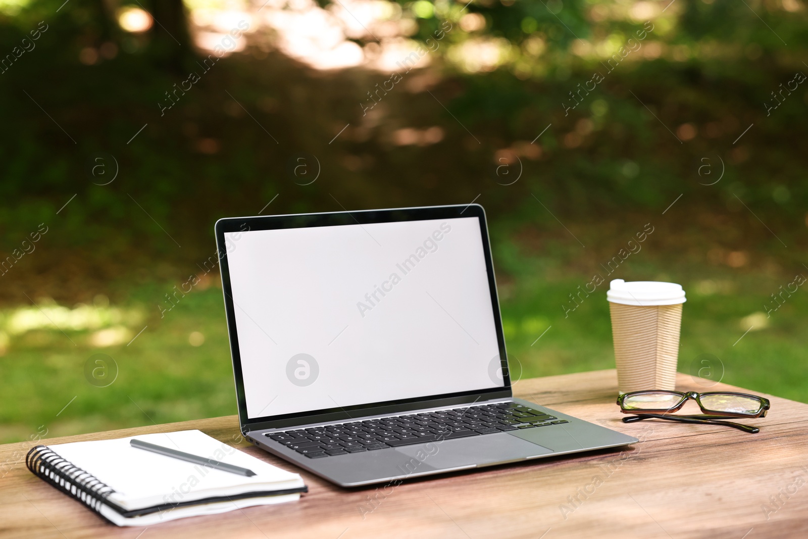 Photo of Laptop, notebook, pen, glasses and paper cup on wooden table outdoors. Remote work