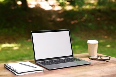 Photo of Laptop, notebook, pen, glasses and paper cup on wooden table outdoors. Remote work