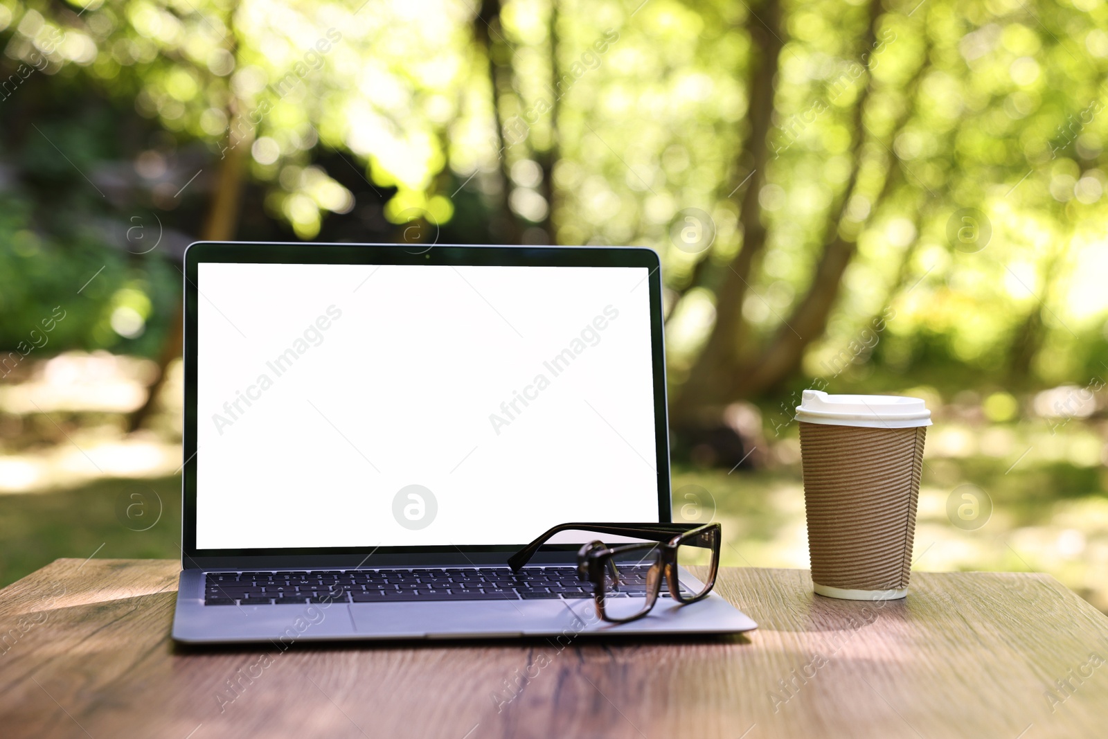 Photo of Laptop, glasses and paper cup on wooden table outdoors. Remote work