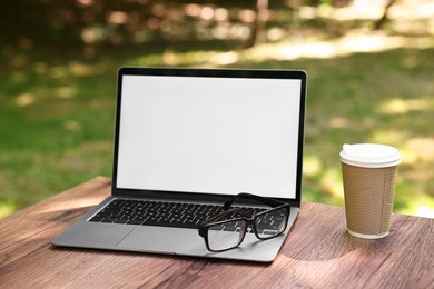 Photo of Laptop, glasses and paper cup on wooden table outdoors. Remote work