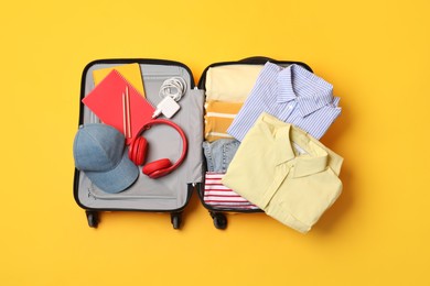 Open suitcase with traveler's belongings on yellow background, top view