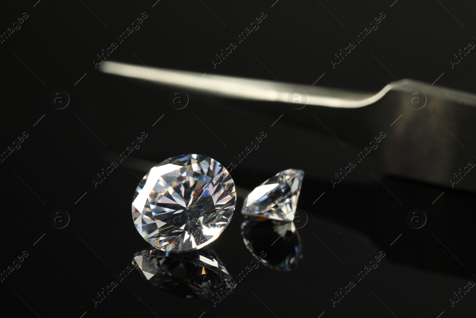 Photo of Beautiful shiny diamonds and tweezers on black mirror surface, closeup