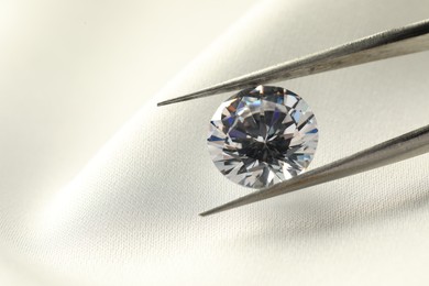 Photo of Tweezers with beautiful shiny diamond on white fabric, closeup. Space for text