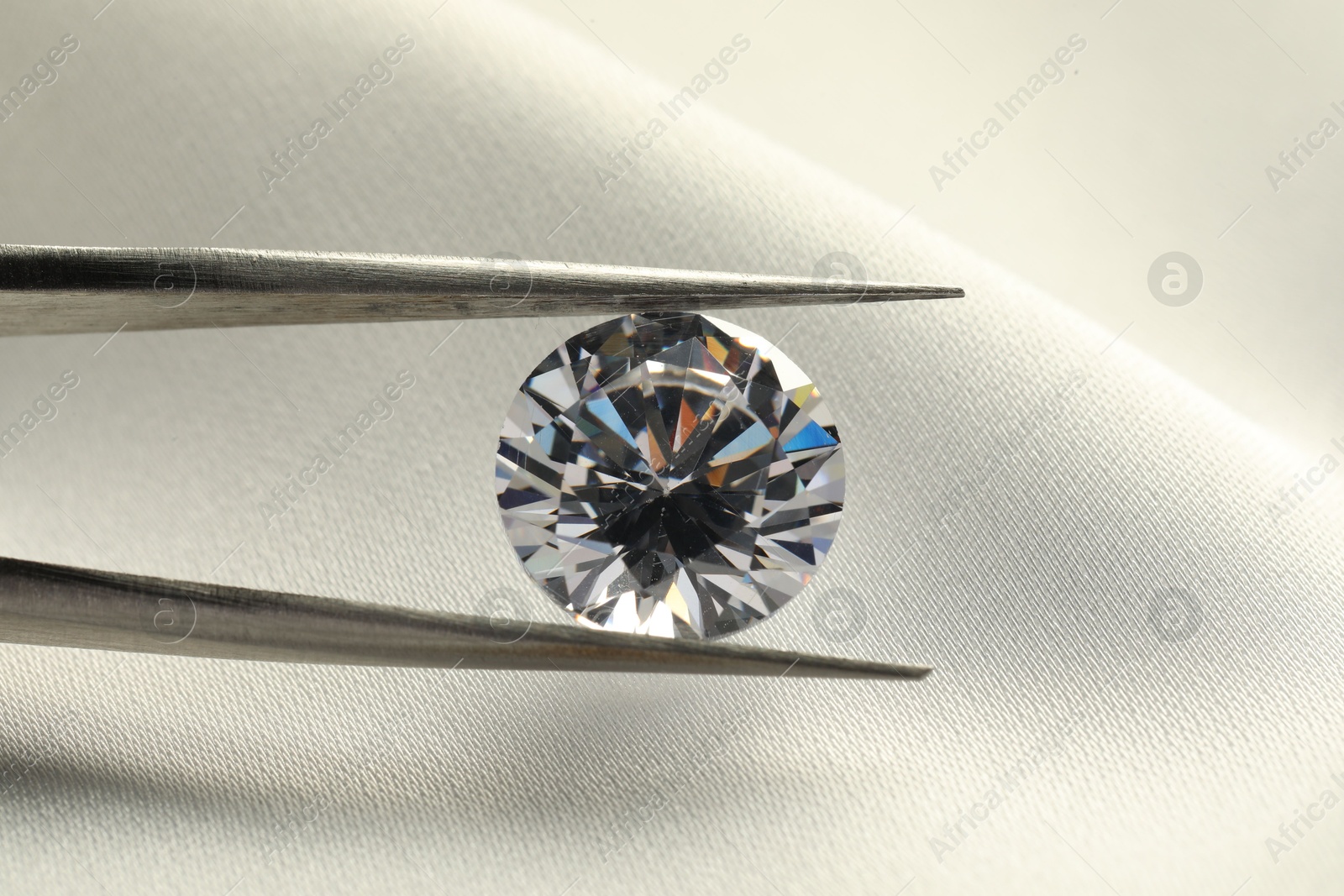 Photo of Tweezers with beautiful shiny diamond on white fabric, closeup