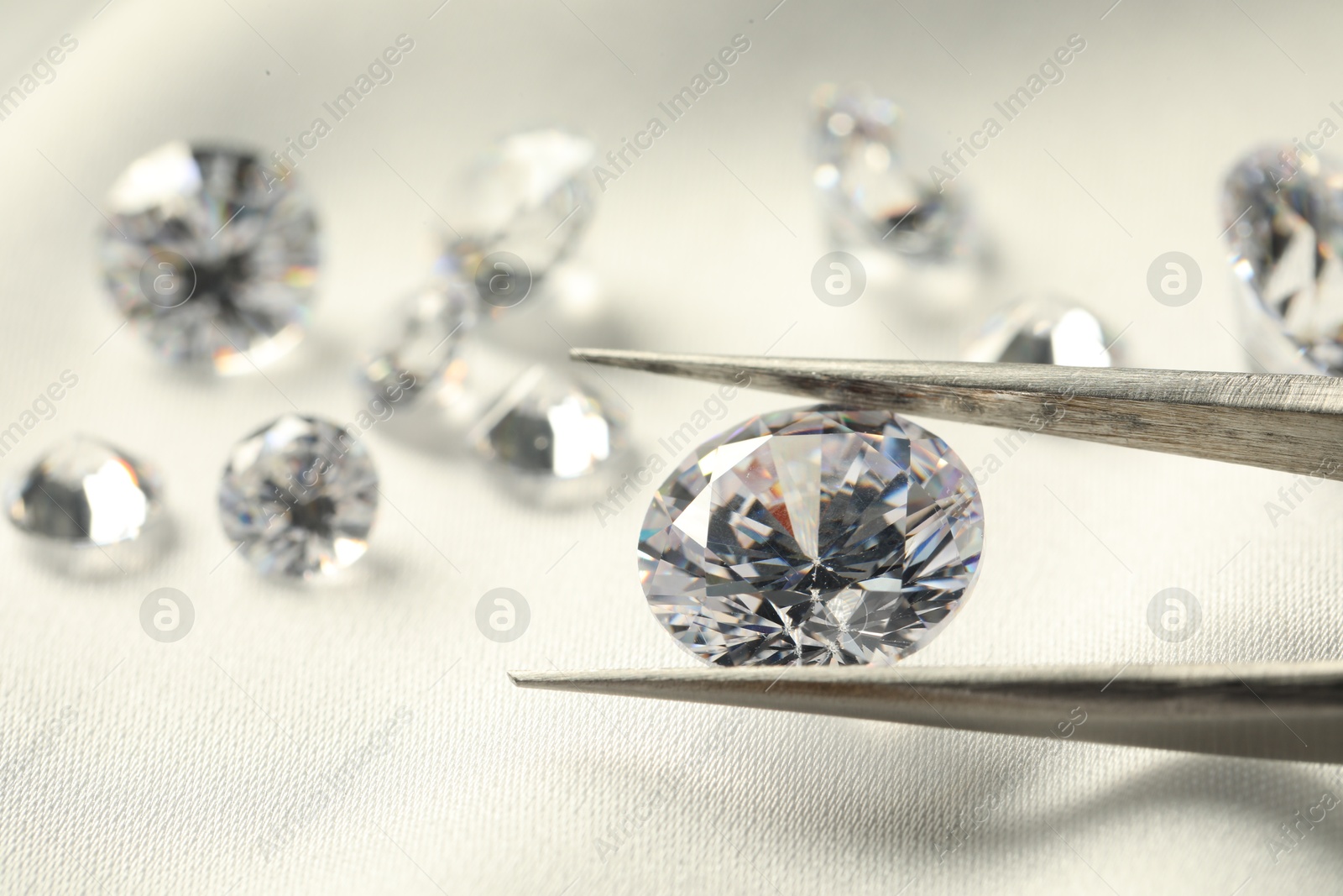 Photo of Tweezers with beautiful shiny diamonds on white fabric, closeup