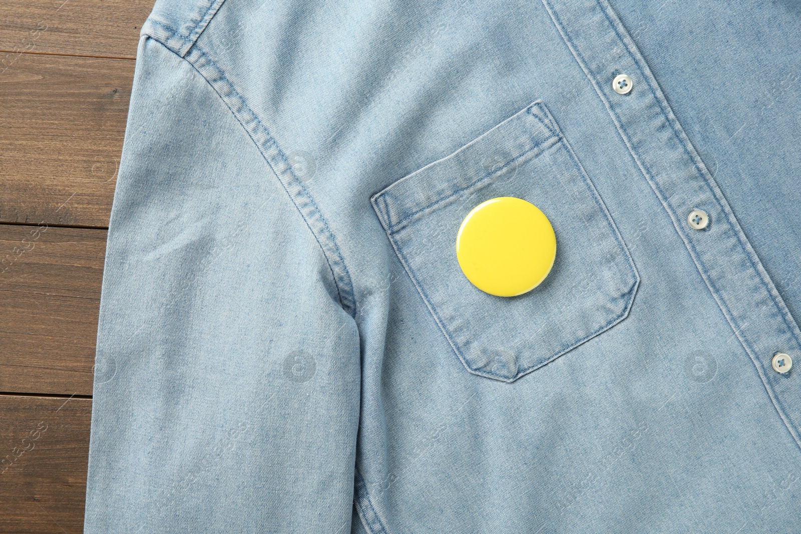 Photo of Denim shirt with blank button badge on wooden background, top view. Mockup for design