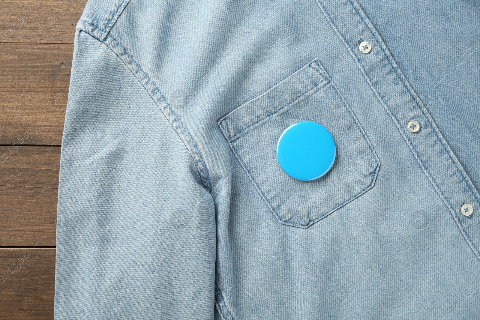 Photo of Denim shirt with blank button badge on wooden background, top view. Mockup for design