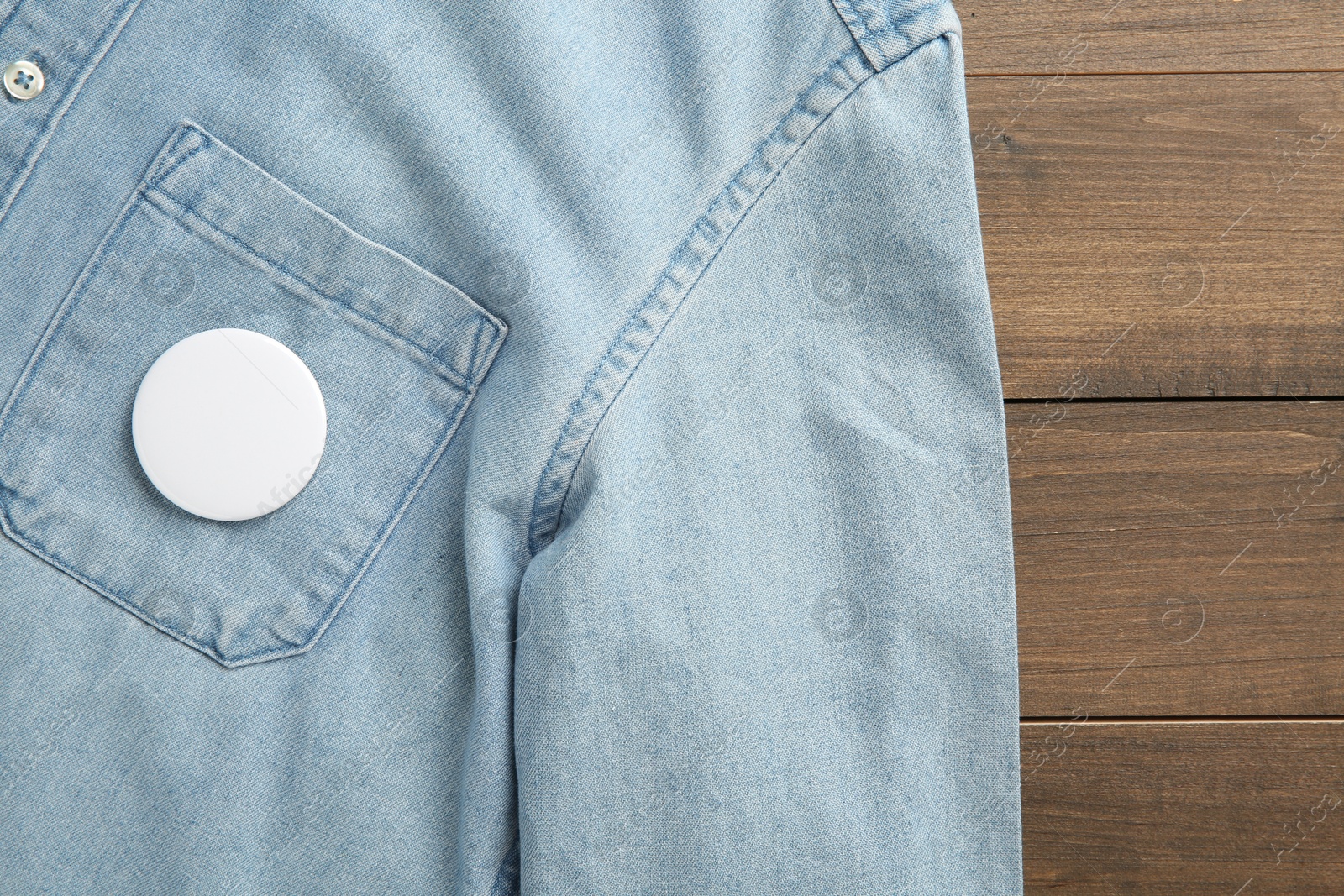 Photo of Denim shirt with blank button badge on wooden background, top view. Mockup for design