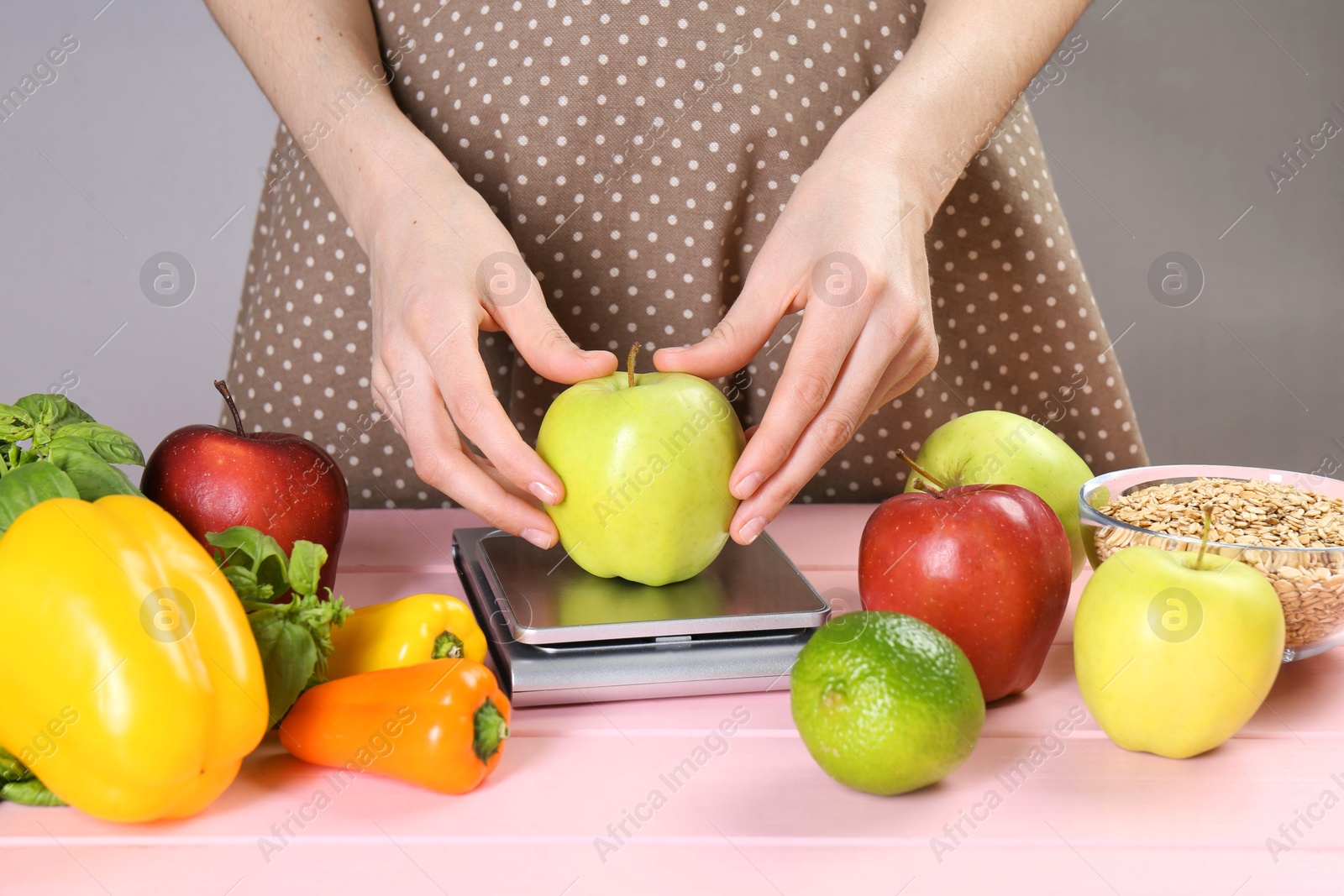 Photo of Woman weighing apple on kitchen scale at pink wooden table, closeup