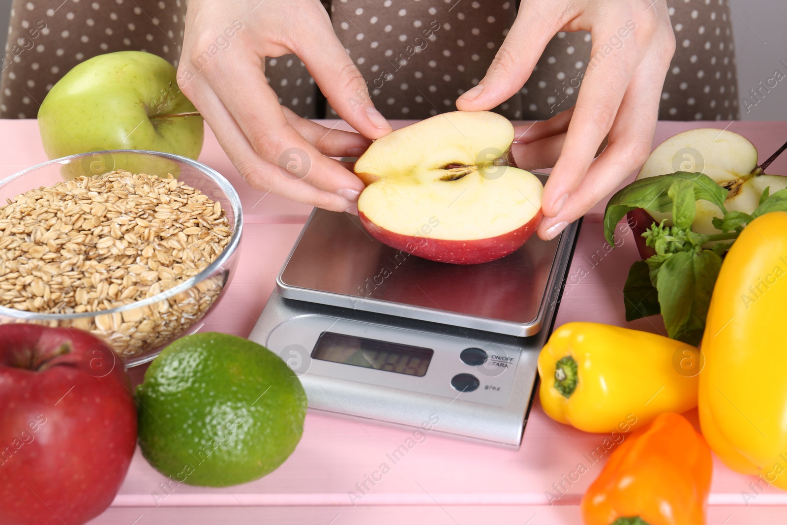 Photo of Woman weighing half of apple on kitchen scale at pink wooden table, closeup