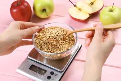 Photo of Woman weighing bowl of oats on kitchen scale at pink wooden table, closeup