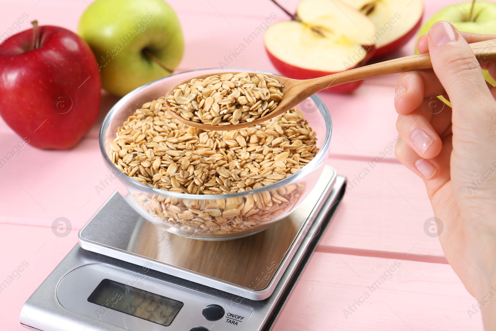 Photo of Woman weighing bowl of oats on kitchen scale at pink wooden table, closeup