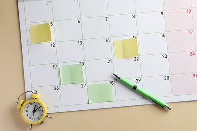 Photo of Timetable. Calendar page with sticky notes, pen and alarm clock on beige background, top view