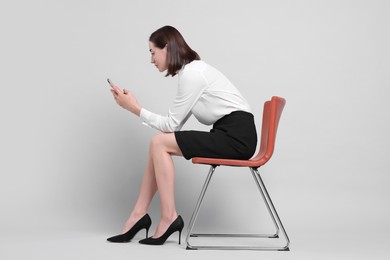 Woman with poor posture sitting on chair and using smartphone against gray background