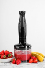 Photo of Hand blender with mixture of ingredients and fresh fruits on white marble table