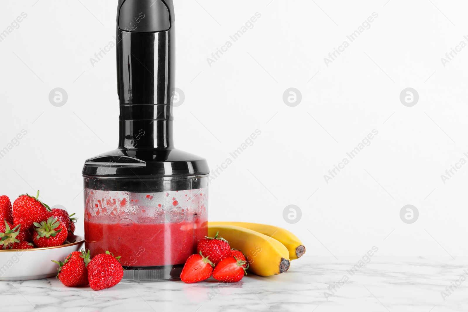 Photo of Hand blender with mixture of ingredients and fresh fruits on white marble table, space for text