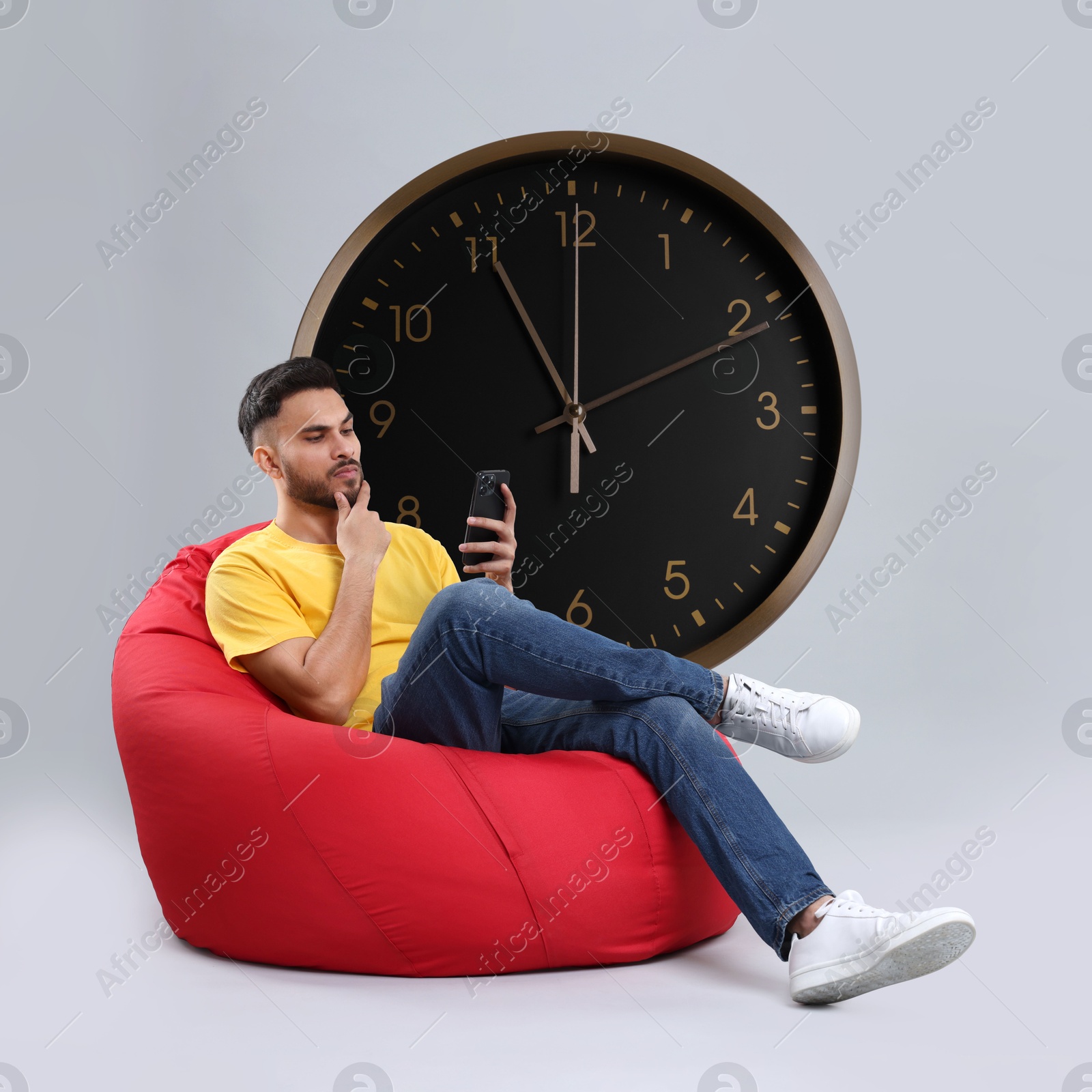 Image of Man with smartphone sitting in beanbag near big clock on grey background