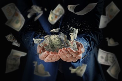 Businessman holding dissolving dollar banknotes in darkness, closeup. Money falling around. Financial crisis