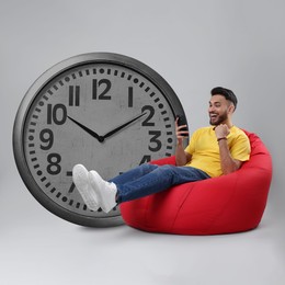 Man with smartphone sitting in beanbag near big clock on grey background