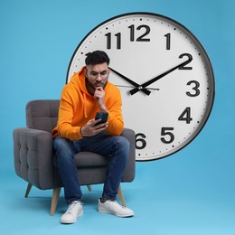 Man with smartphone sitting in armchair near big clock on light blue background