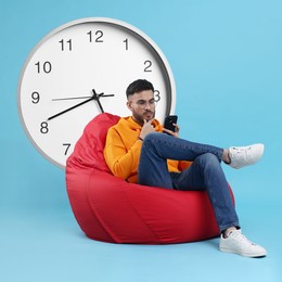 Man with smartphone sitting in beanbag near big clock on light blue background