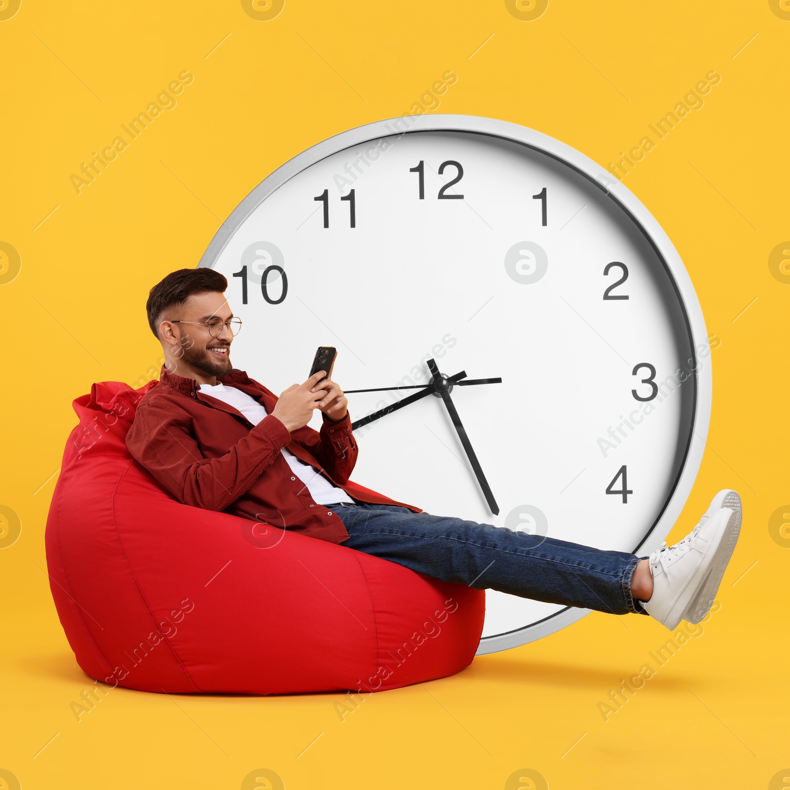 Image of Man with smartphone sitting in beanbag near big clock on orange background