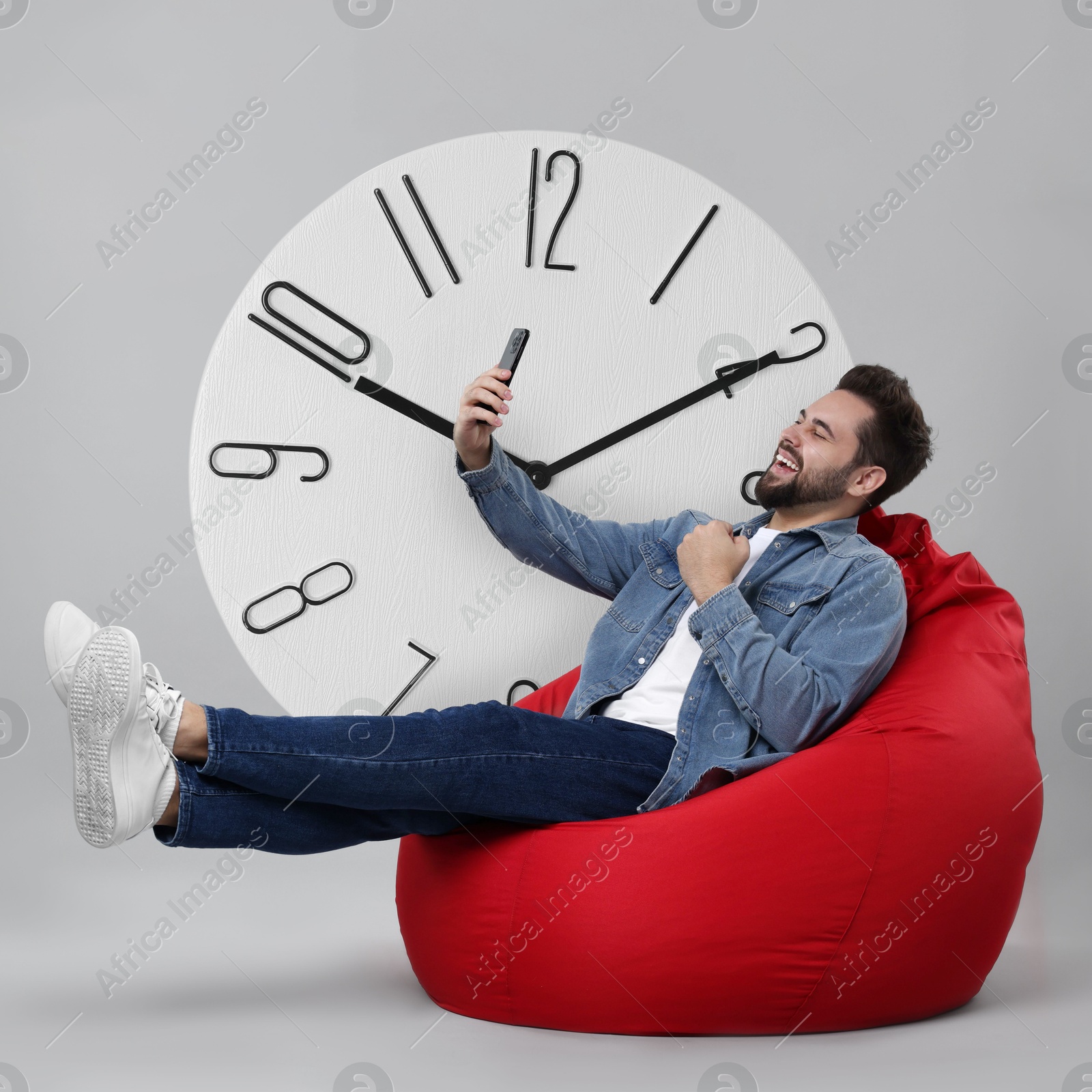 Image of Man with smartphone sitting in beanbag near big clock on grey background