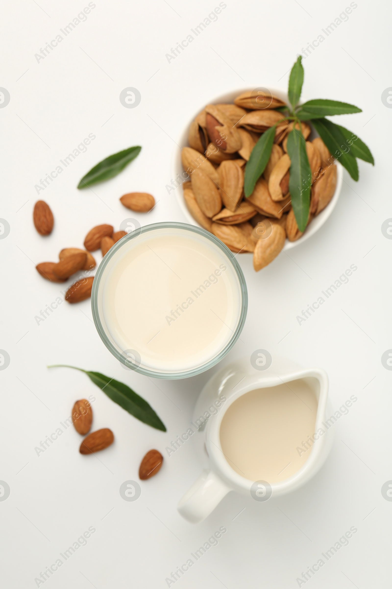 Photo of Fresh almond milk in glass, nuts, green leaves and pitcher on white background, flat lay