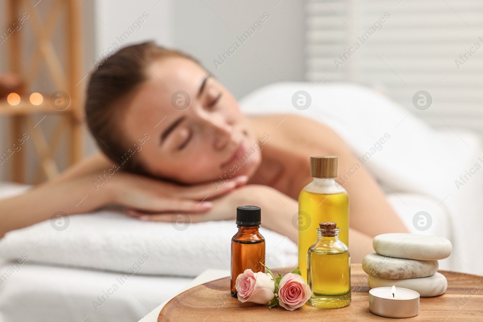 Photo of Aromatherapy. Woman relaxing on massage couch in spa salon, focus on bottles of essential oils, burning candle, stones and rose flowers