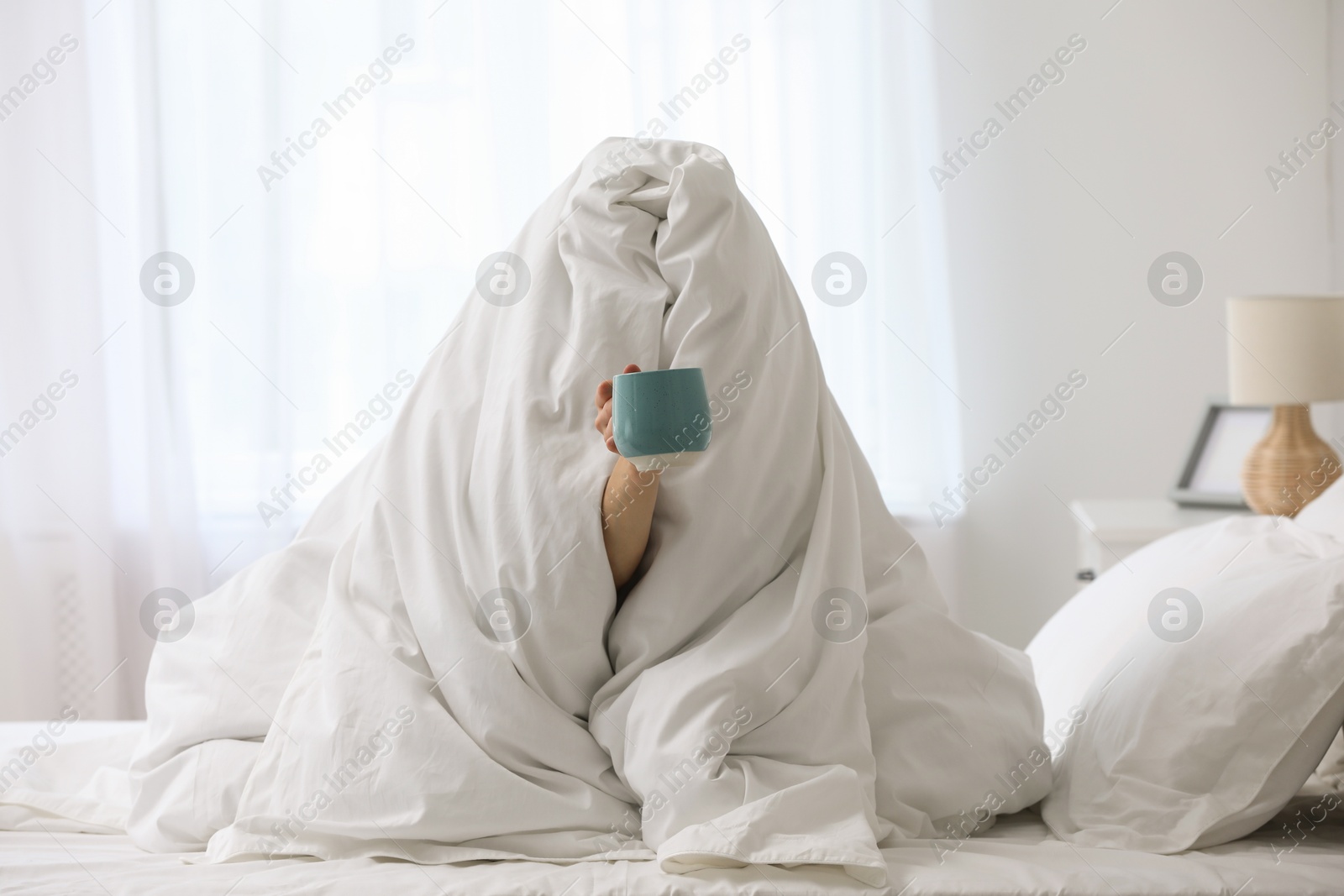 Photo of Woman wrapping in blanket sitting with cup of coffee on bed. Good morning